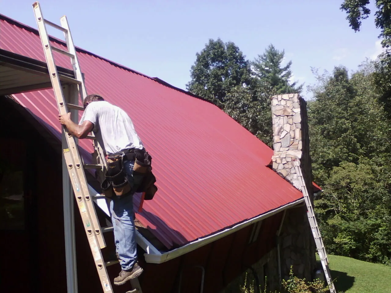 Roofer on a ladder beside a red metal roof and stone chimney — Soffit & Fascia Repair by Roofing Brothers in Bridgeton